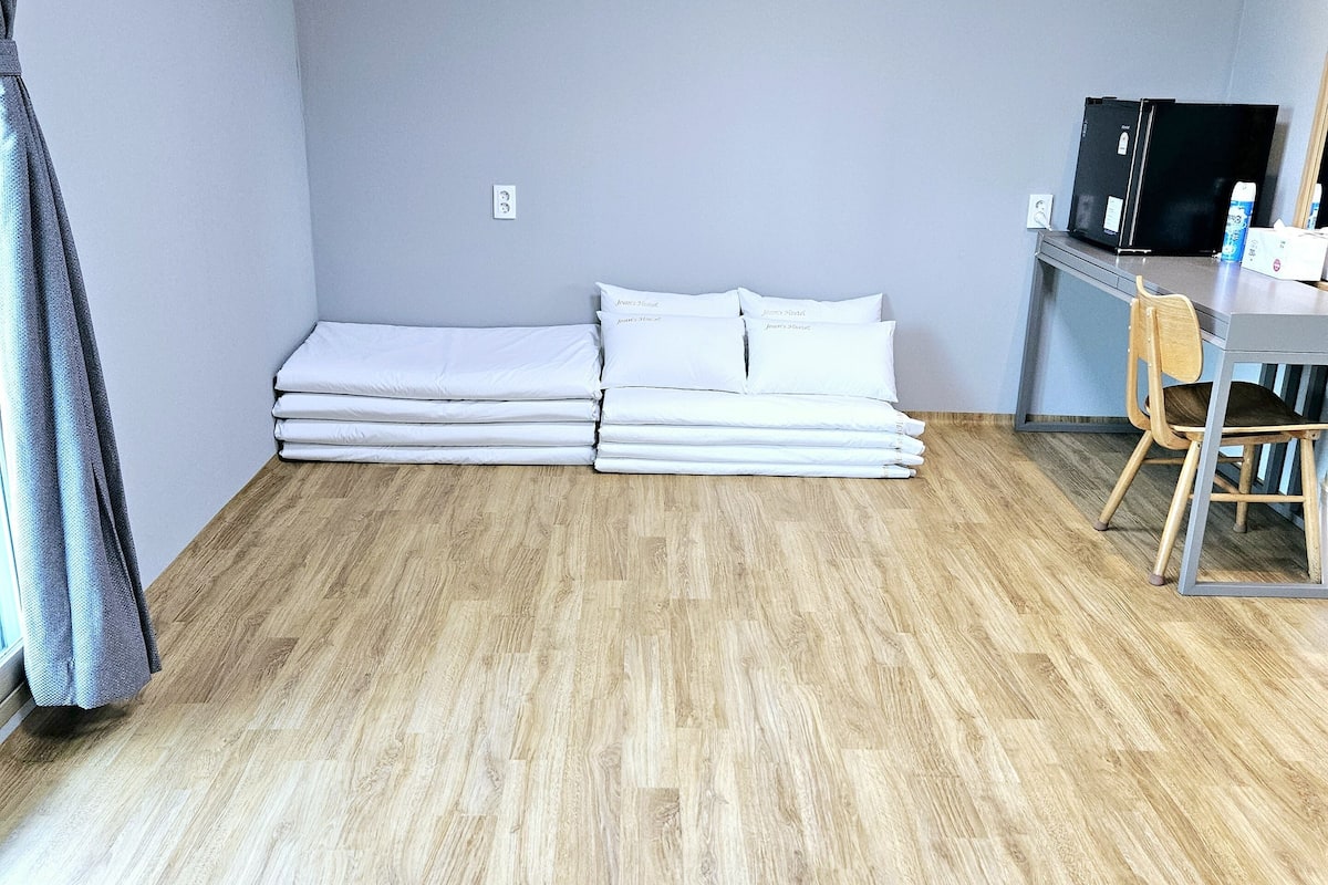 Una habitación con piso de madera, cama con ropa de cama blanca, escritorio con silla y refrigerador negro.