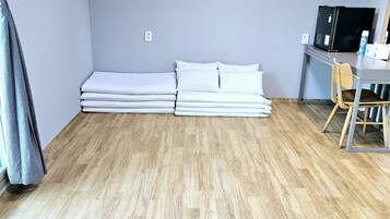 Una habitación con piso de madera, cama con ropa de cama blanca, escritorio con silla y refrigerador negro.