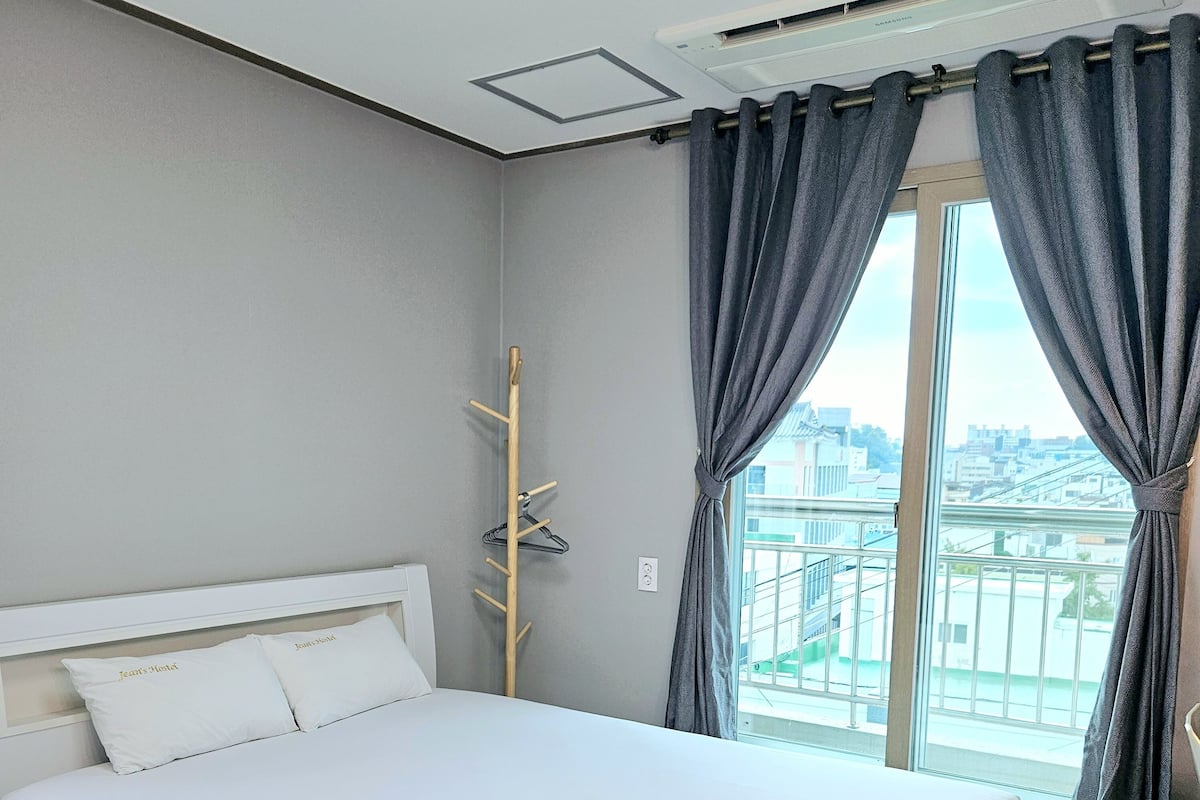 Una habitación de hotel con una cama, un perchero de madera y vistas a la ciudad a través de grandes ventanales.