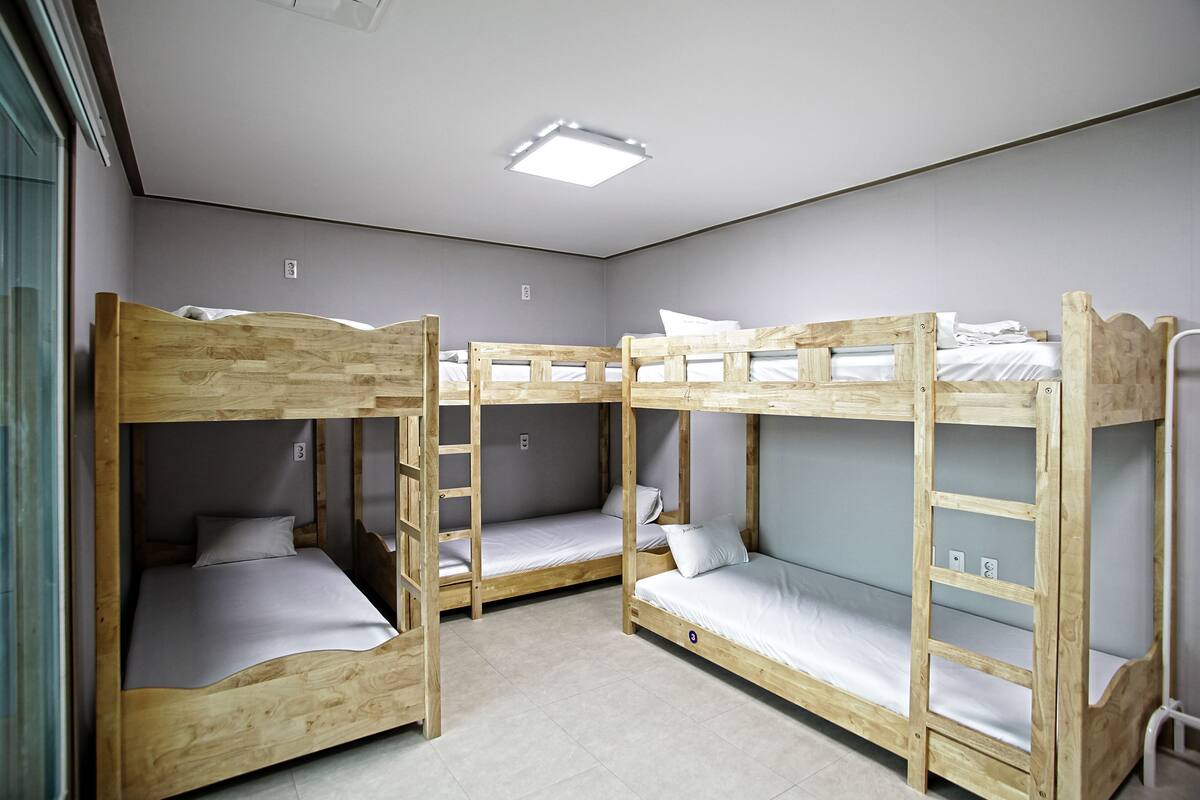Una habitación con tres literas, cada una con estructura de madera y ropa de cama blanca. Hay una lámpara en el techo sobre las camas.