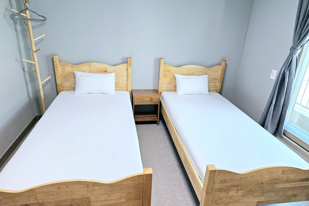 Habitación doble con dos camas de madera, una mesita de noche de madera, un perchero y una ventana con cortinas.