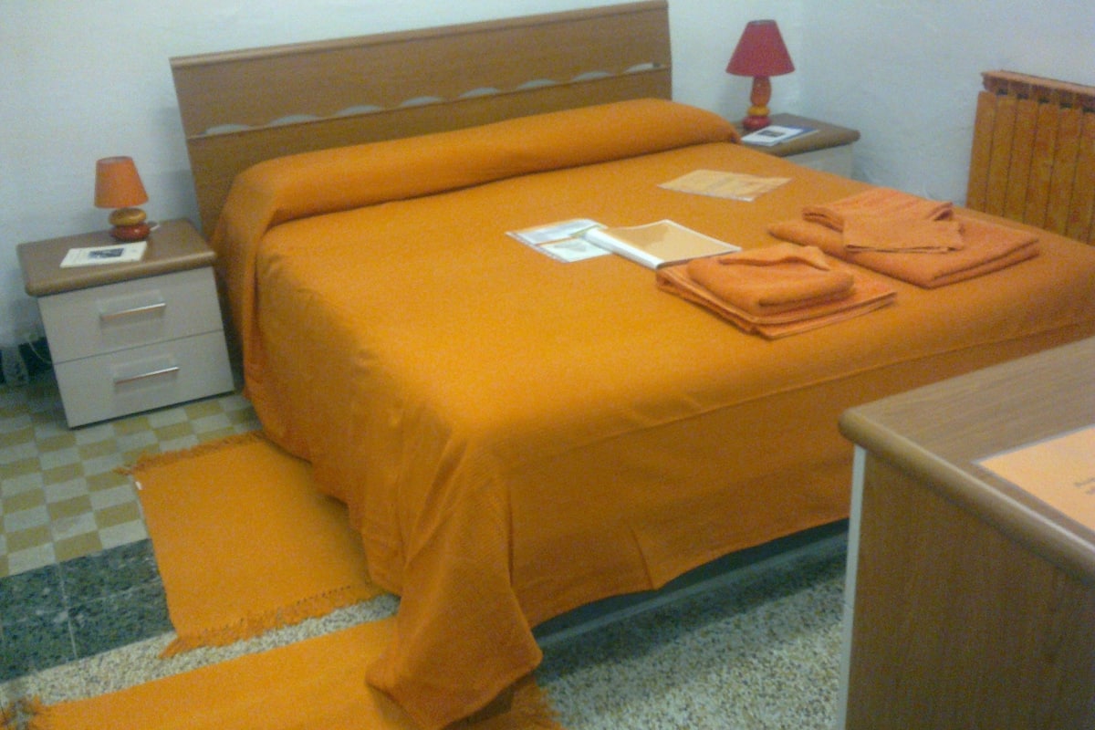 A neatly made bed with an orange bedspread, a bedside table with a lamp, and a small drawer unit.