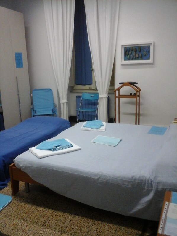 A hotel room with two beds, a blue chair, a wooden chair, and a window with white curtains.