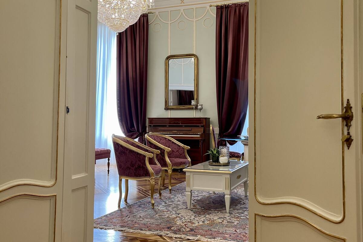 An elegantly furnished room with a grand chandelier, a piano, and plush red armchairs.