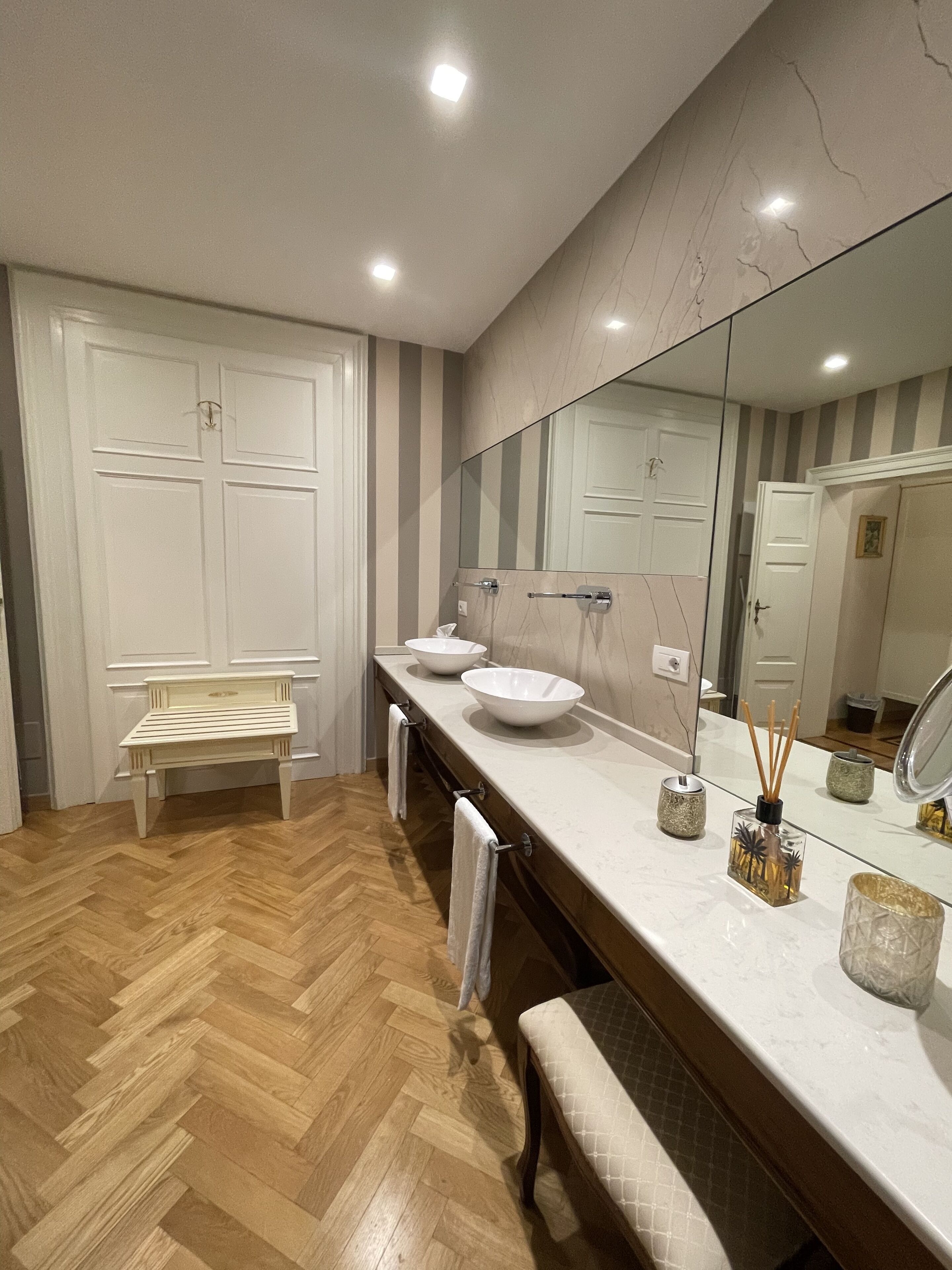 A modern bathroom with a wooden herringbone floor, a bench, two sinks, and a large mirror.