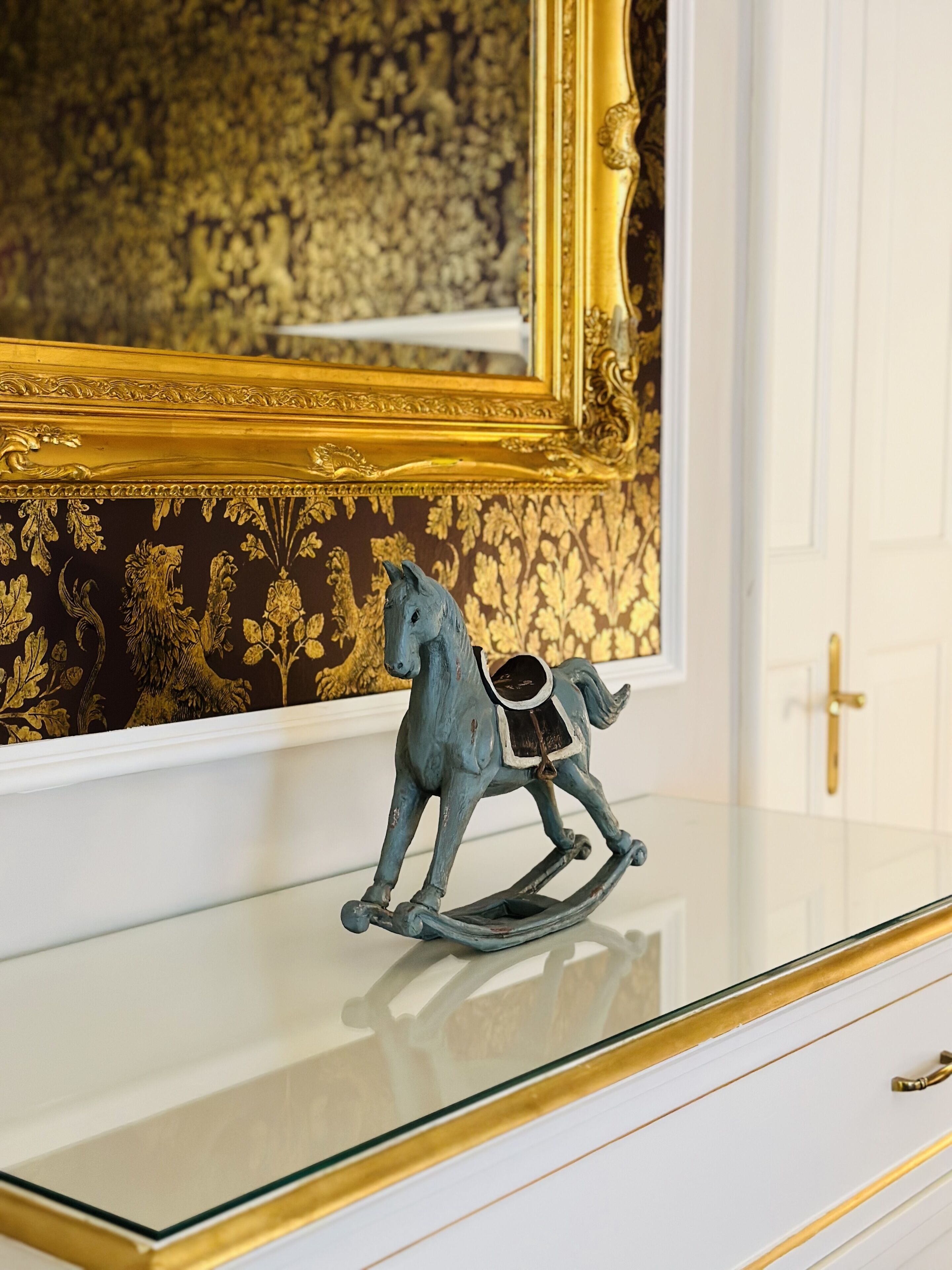 Una habitación con un tocador de superficie de vidrio, un caballito de madera decorativo, un espejo con marco dorado y un papel tapiz estampado.