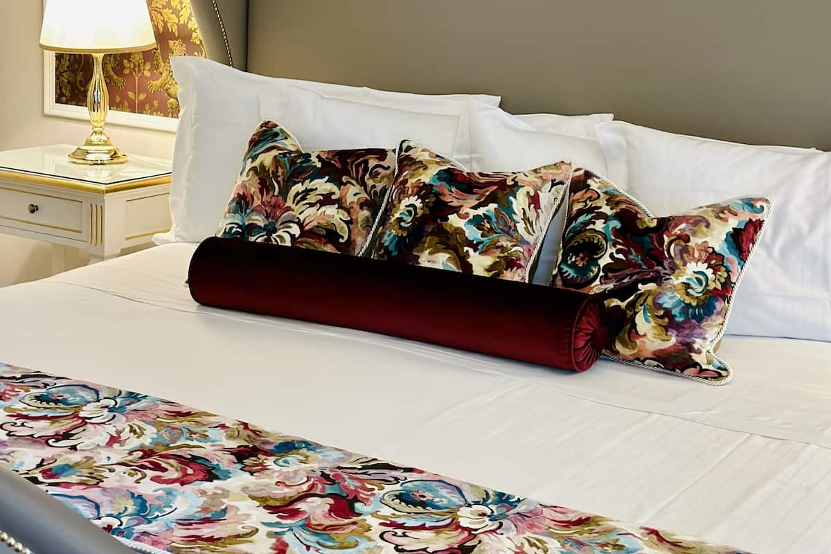 A neatly made bed with a floral bedspread and two decorative pillows, a bedside lamp, and a wall with a patterned wallpaper.