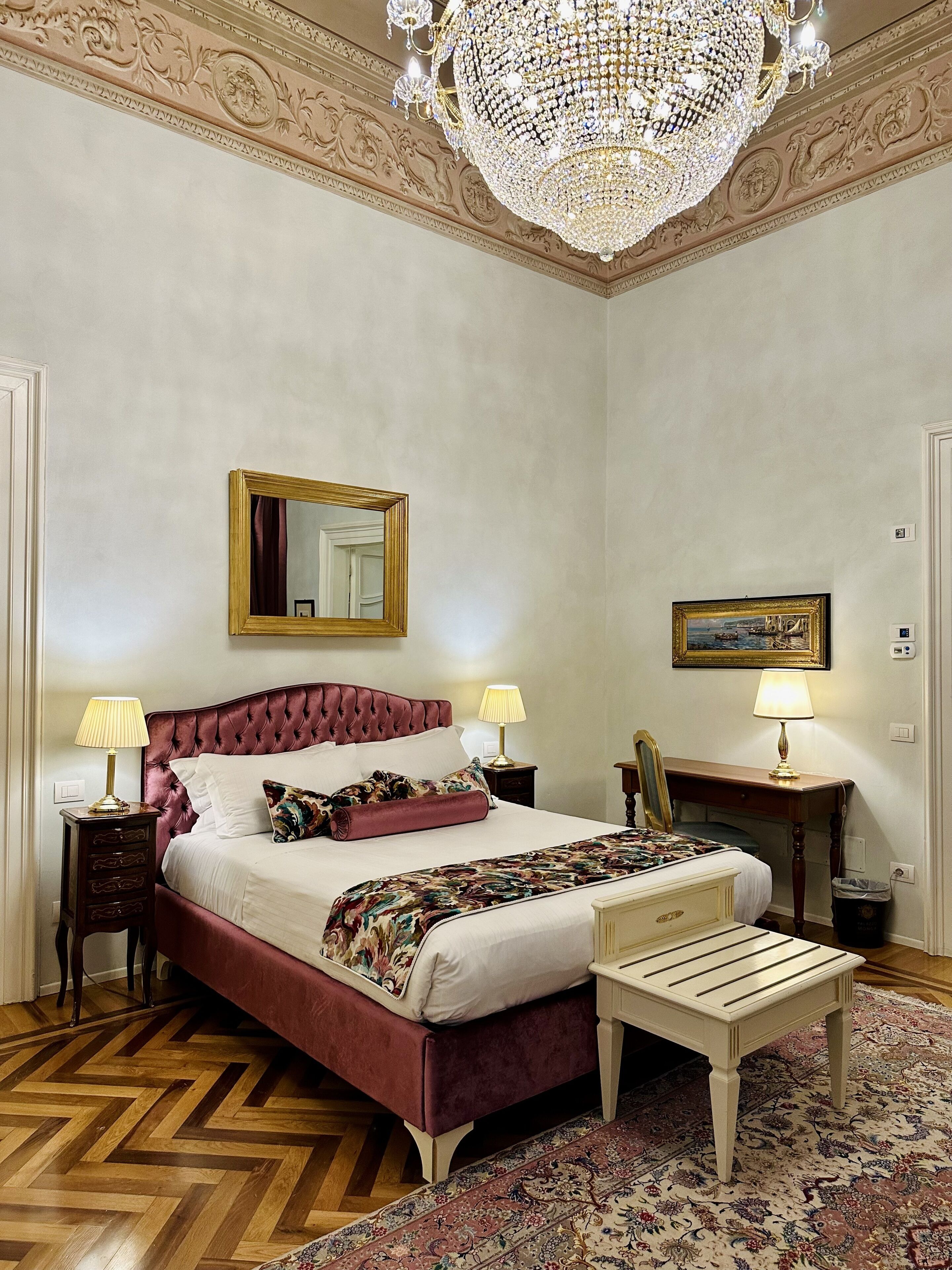 A bedroom with a large bed, two bedside tables with lamps, a mirror, a picture frame, and a chandelier.