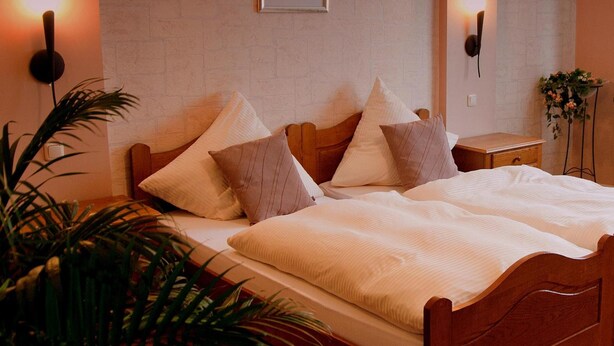 A neatly made bed with a wooden frame, a bedside table with a lamp, and a wall-mounted light fixture. - Hotel zum Schnackel (Wiesbaden)
