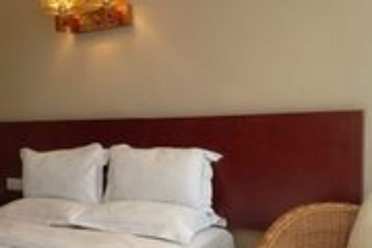 A hotel room with a bed, a headboard, a wicker chair, and a wall-mounted light fixture.