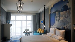 A hotel room with a large bed, a blue armchair, a small table, and a painting of a coastal scene. - Huaqing Aegean Inter Hot Spring Resort (Xi'an)