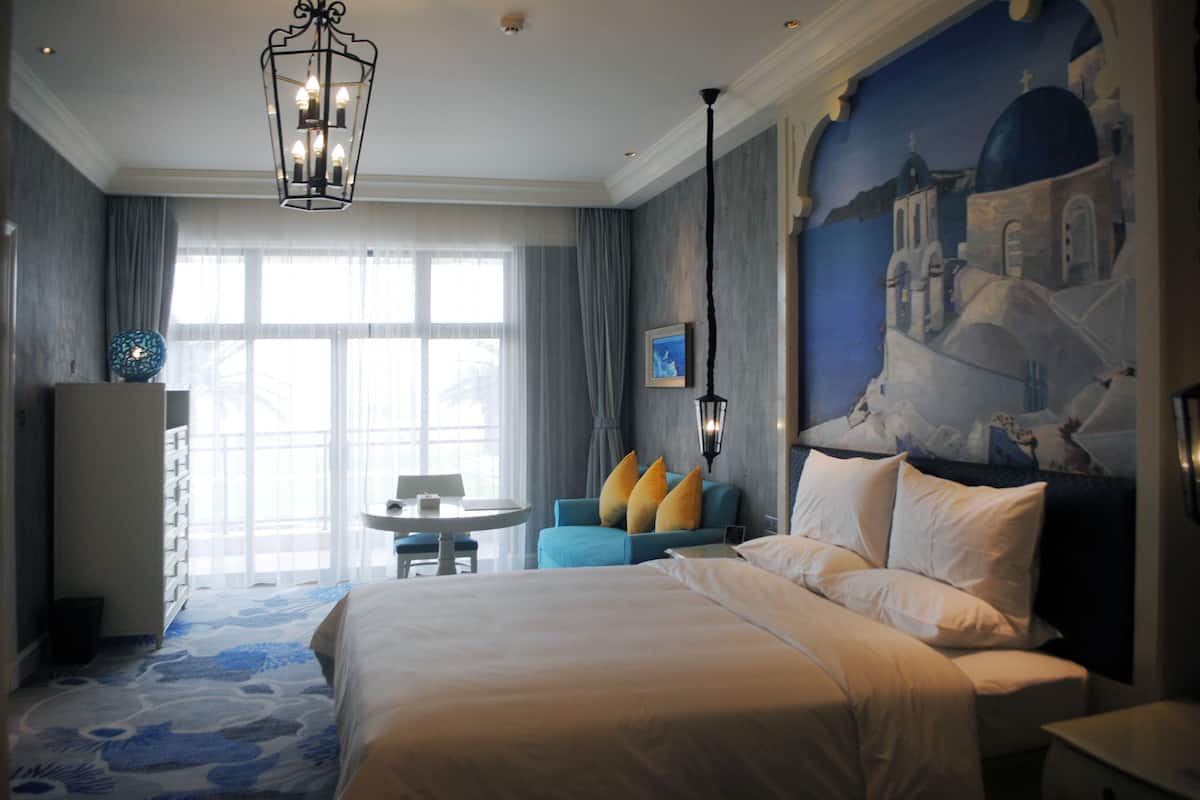 A hotel room with a large bed, a blue armchair, a small table, and a painting of a coastal scene.