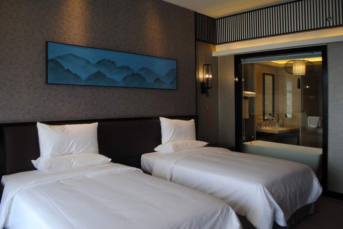 A hotel room with two beds, a large painting on the wall, and a bathroom visible through a glass door.