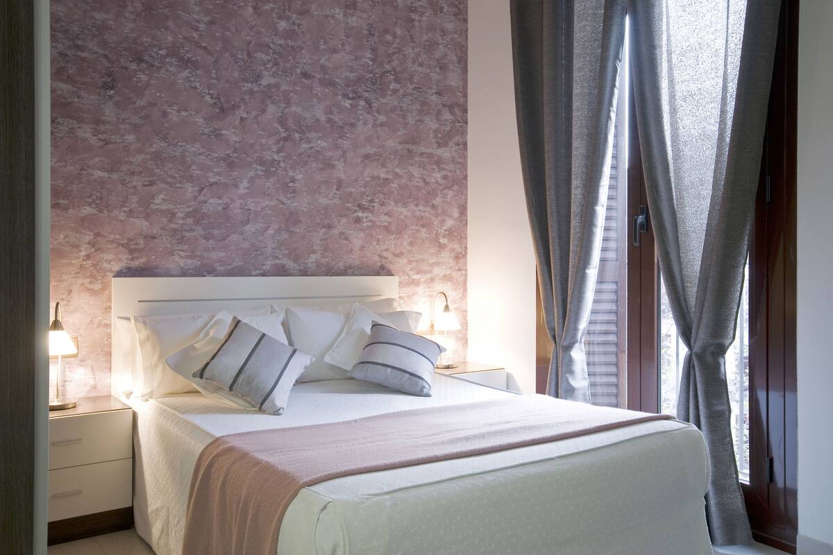 A neatly made bed with pillows and a blanket, a bedside table with a lamp, and a window with curtains.