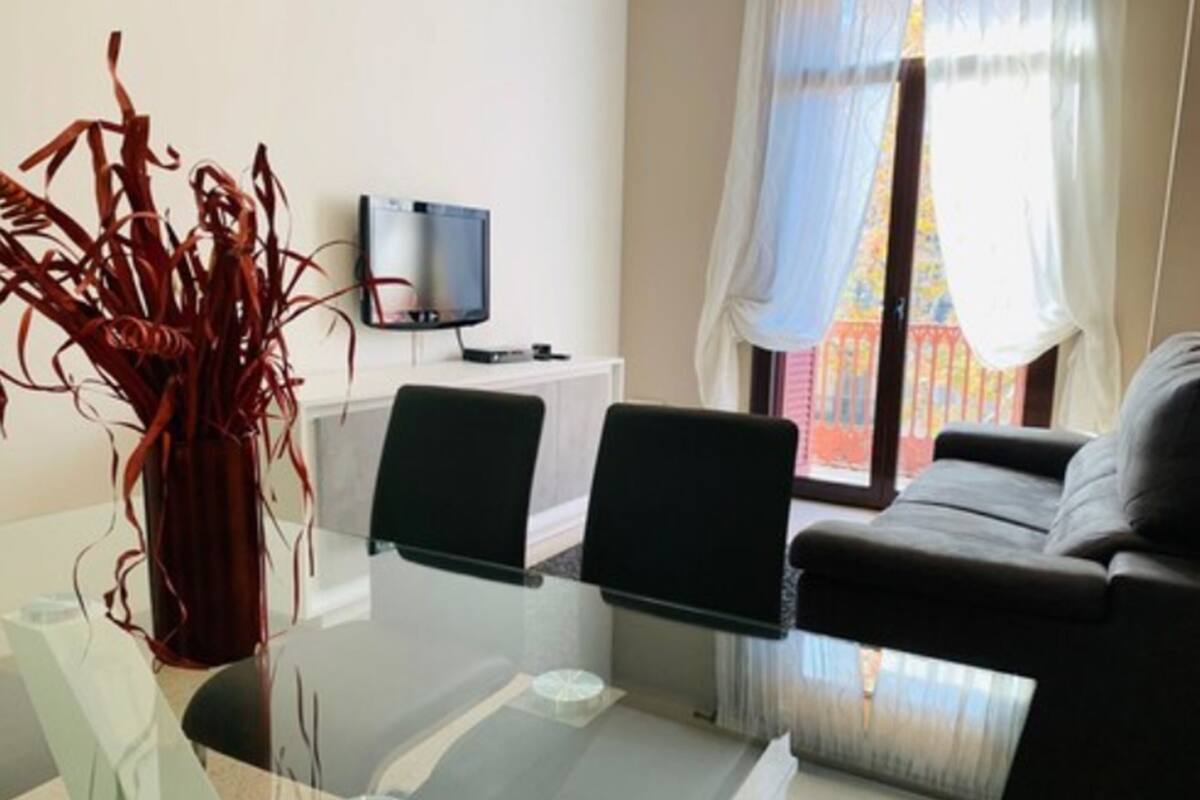 A modern living room with a glass table, two black chairs, a sofa, a TV on a stand, and a window with sheer curtains.