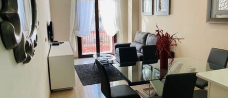 A modern living room with a glass dining table, black chairs, a grey sofa, and a red decorative plant.