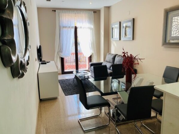 A modern living room with a glass dining table, black chairs, a grey sofa, and a red decorative plant.