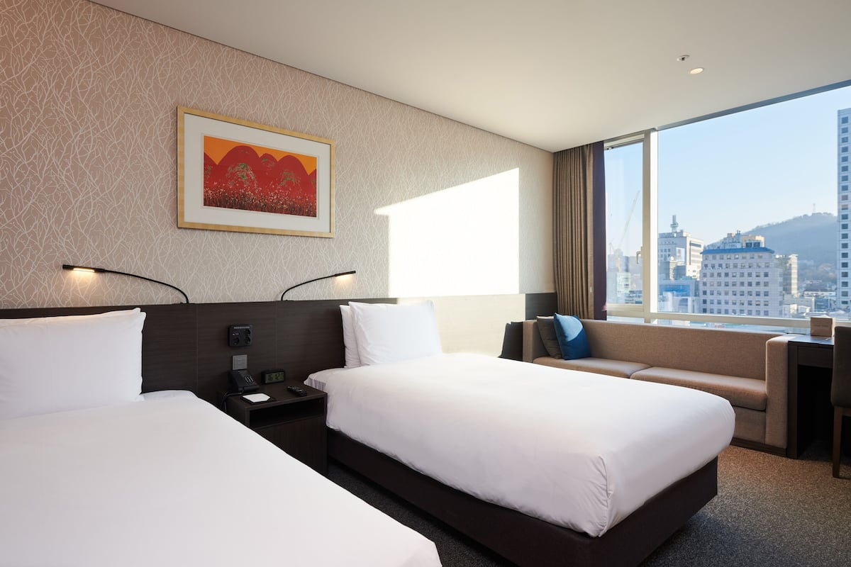 A hotel room with two beds, a desk, a chair, and a large window offering a city view.