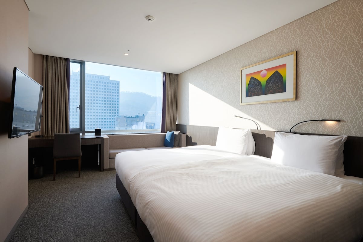A hotel room with a large bed, a desk with a chair, a TV, and a window with a city view.