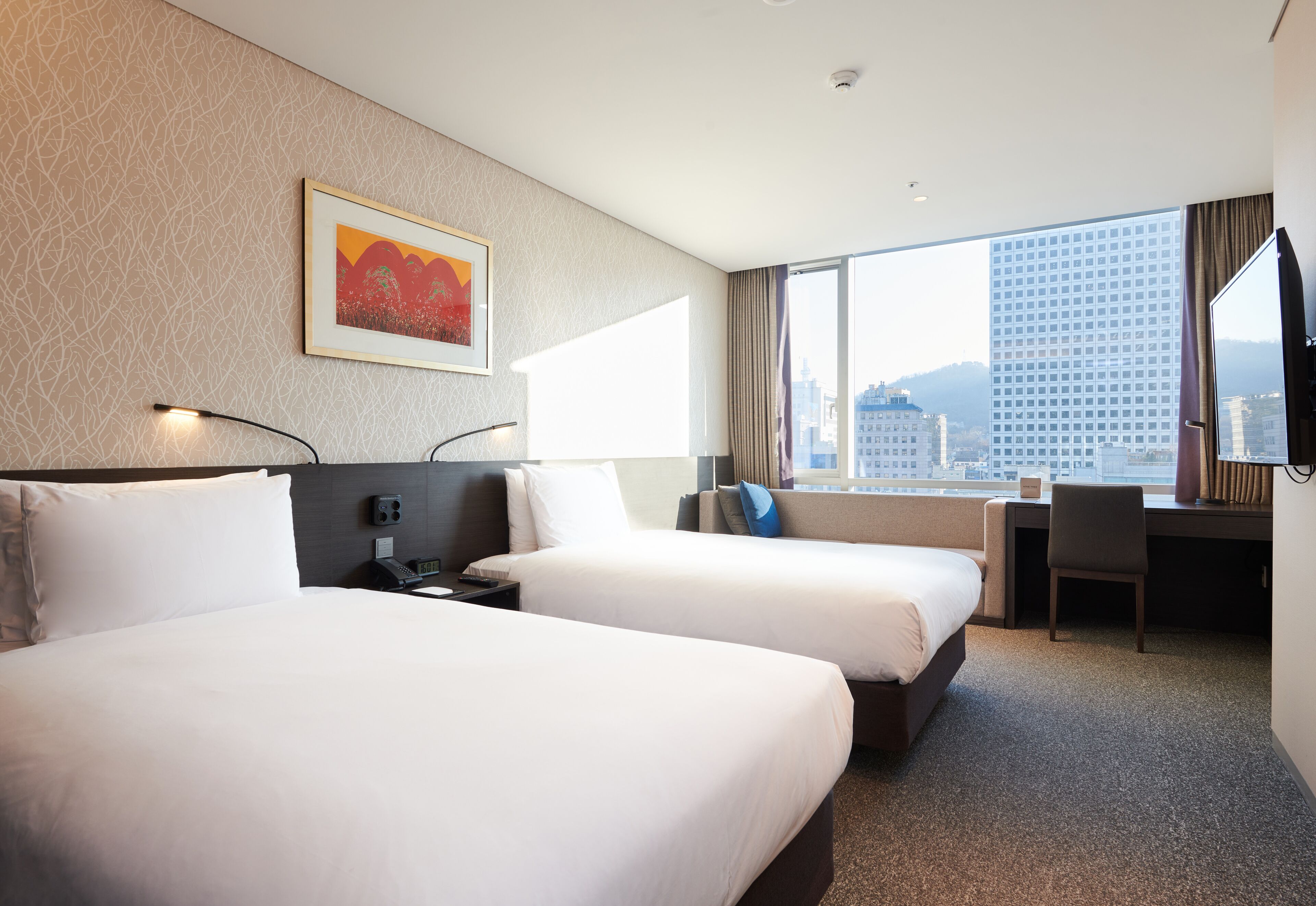 Ein Hotelzimmer mit zwei Betten, einem Schreibtisch und einem großen Fenster mit Blick auf die Stadt.