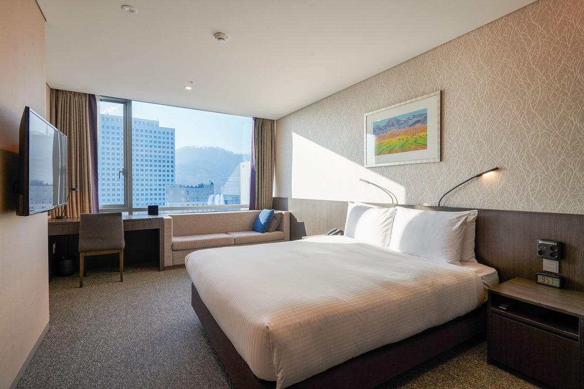 A hotel room with a large bed, a desk, a chair, a TV, and a window with a city view.
