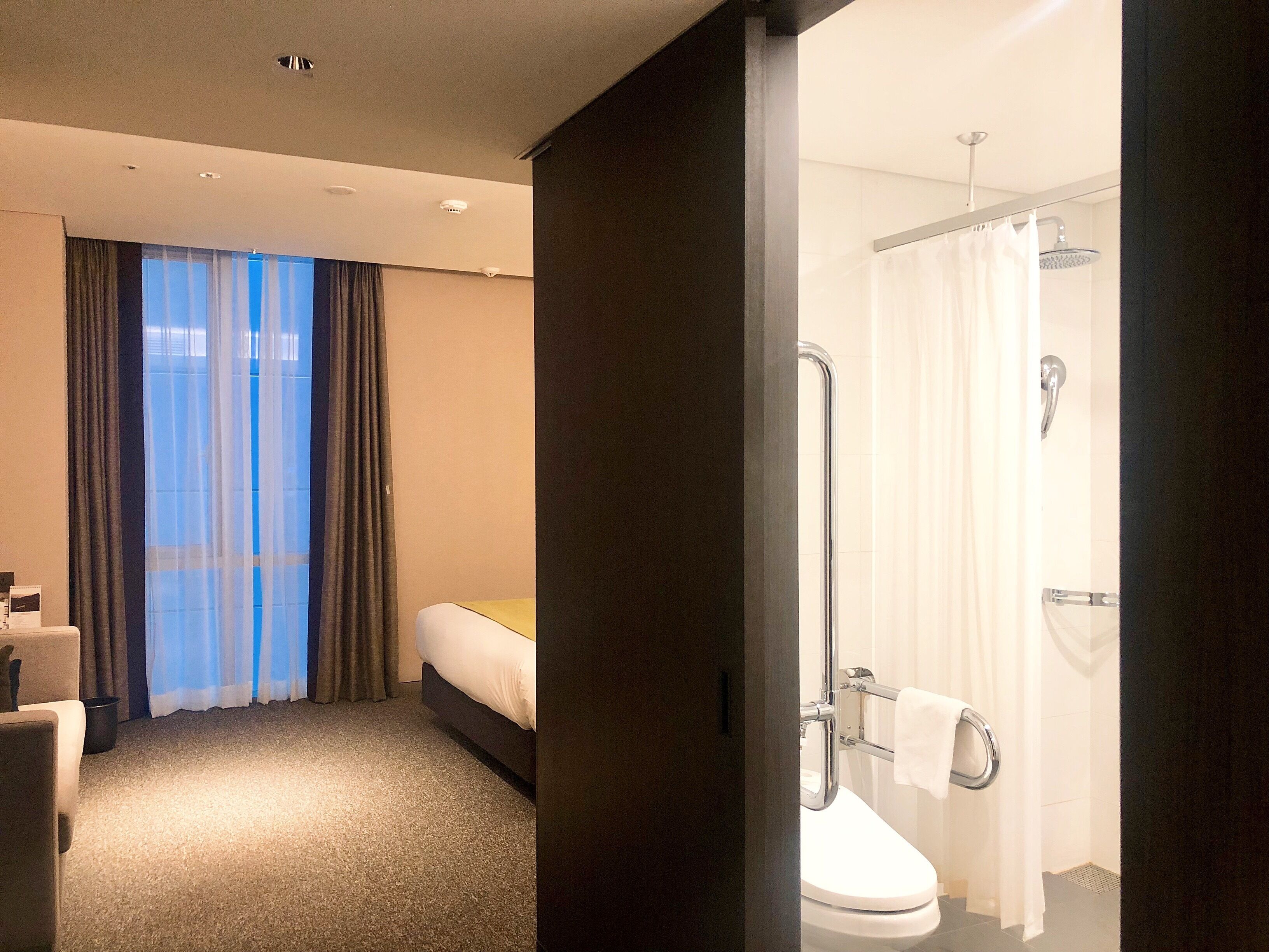 A hotel room with a bed, a bathroom with a shower, and a view of the outdoors through a window.