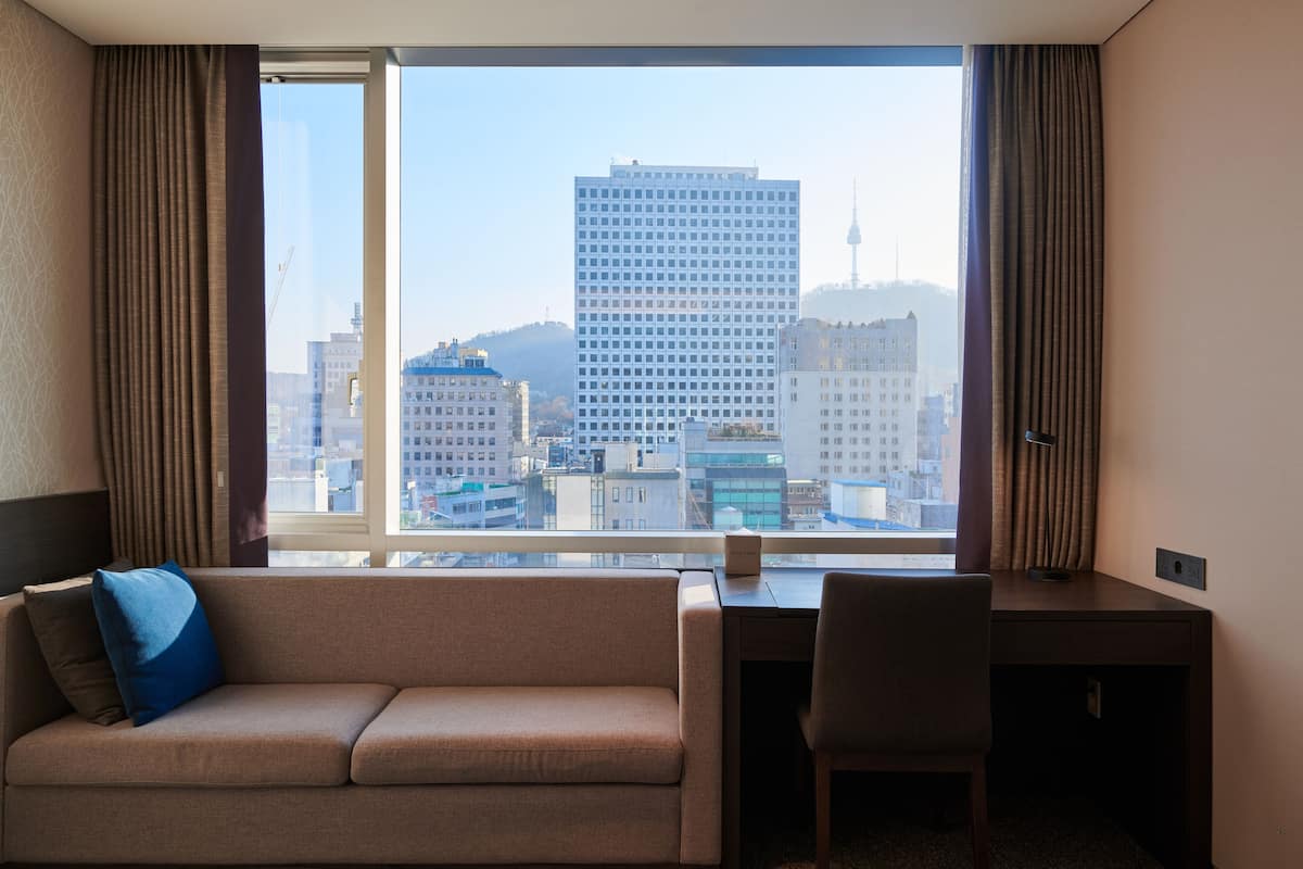 A hotel room with a large window, a sofa, a desk, and a chair.