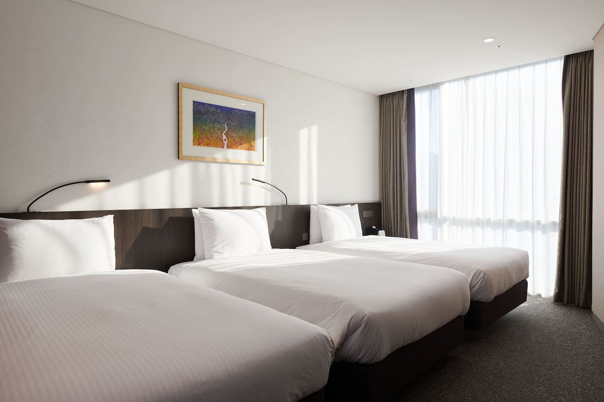 A hotel room with three beds, a large window with curtains, and a framed picture on the wall.