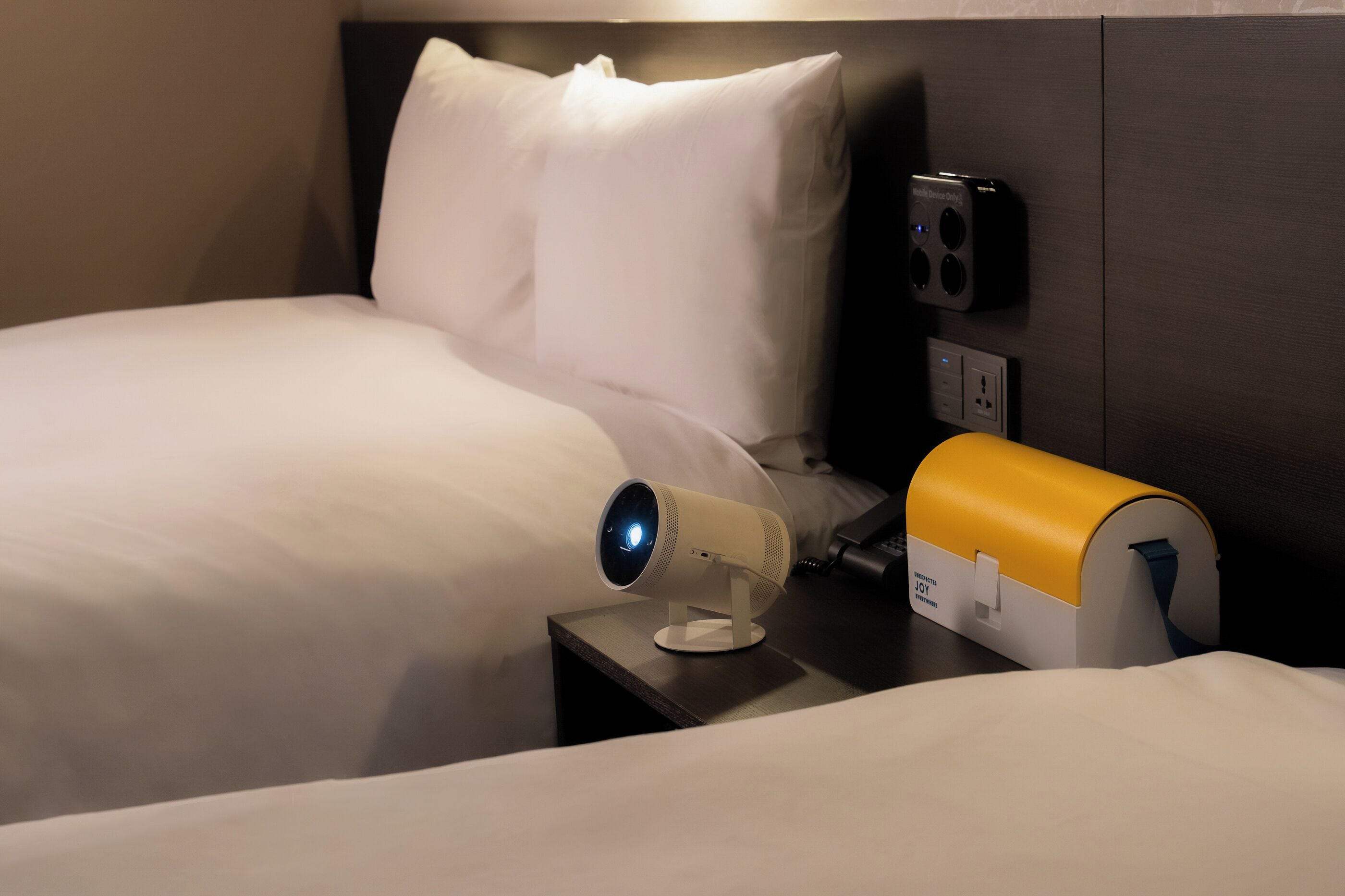 A hotel room with a bed, a nightstand, a white lamp, and a device on the nightstand.