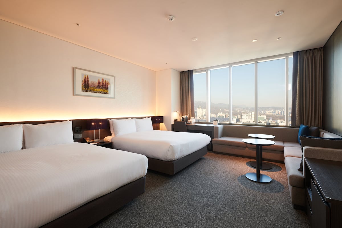 A hotel room with two beds, a sofa, a small table, and a large window offering a city view.