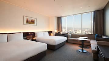 Quarto de hotel com duas camas, um sofá, uma mesinha e uma janela grande com vista para a cidade.