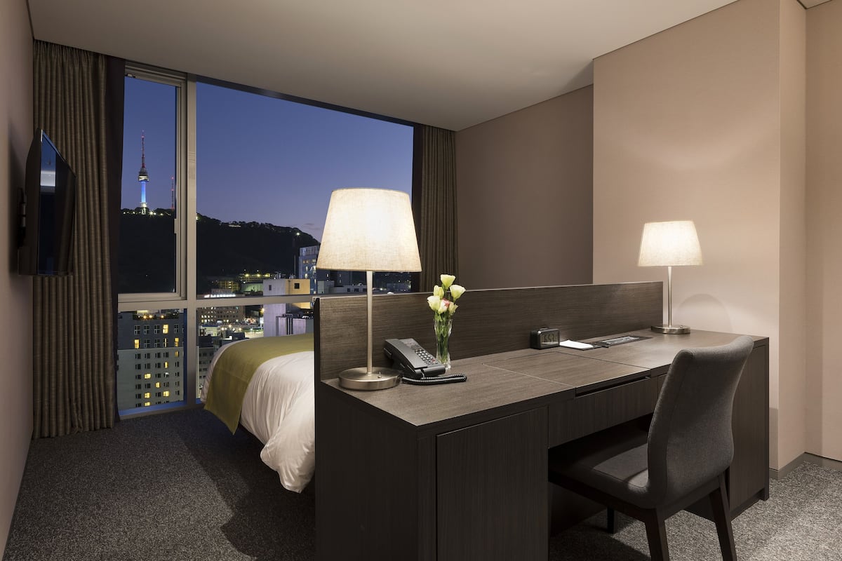 A hotel room with a desk, chair, bed, and a large window offering a city view.
