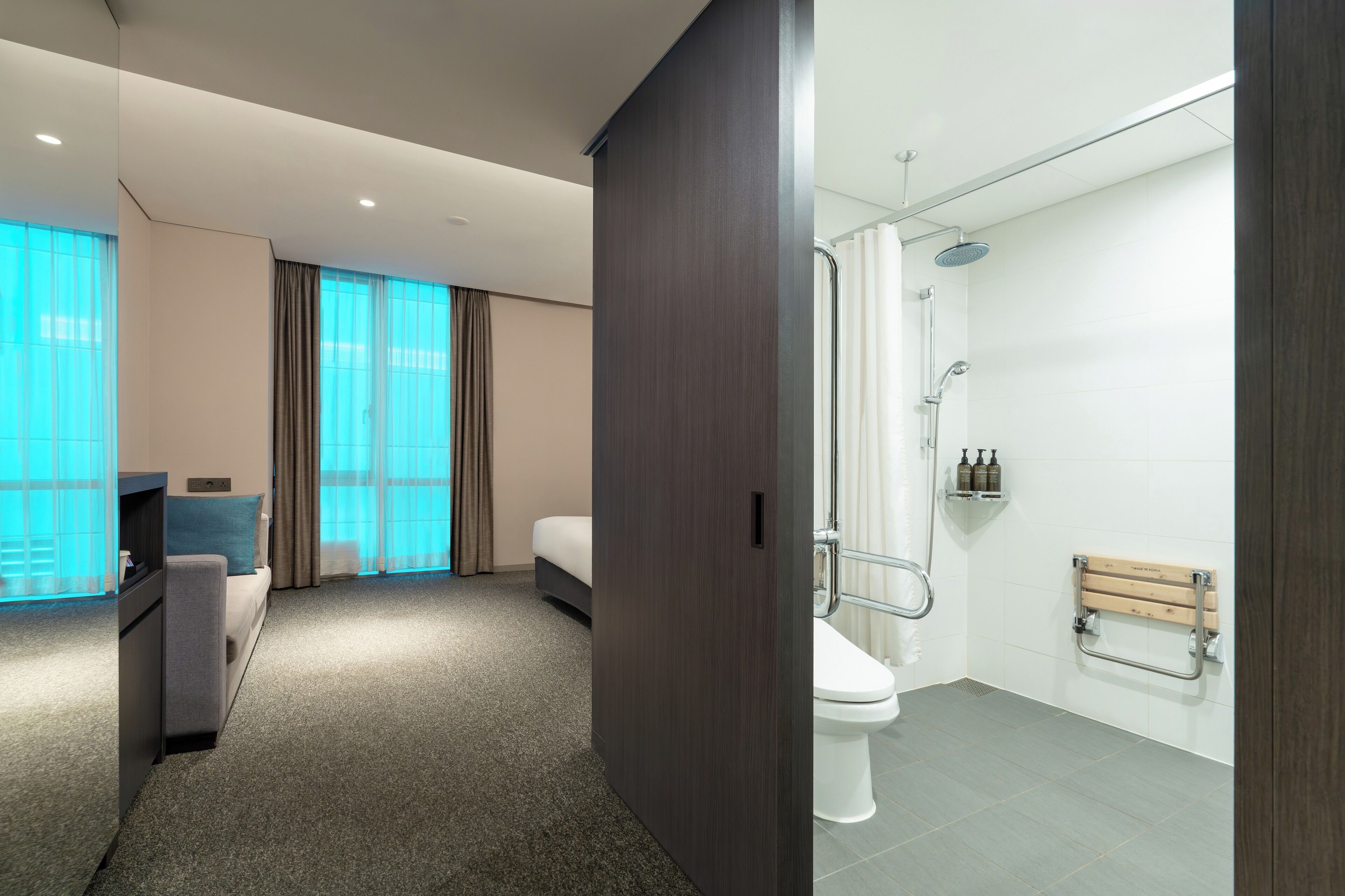 A modern hotel room with a sofa, a TV, a large window with curtains, and a separate bathroom with a shower, toilet, and towel rack.