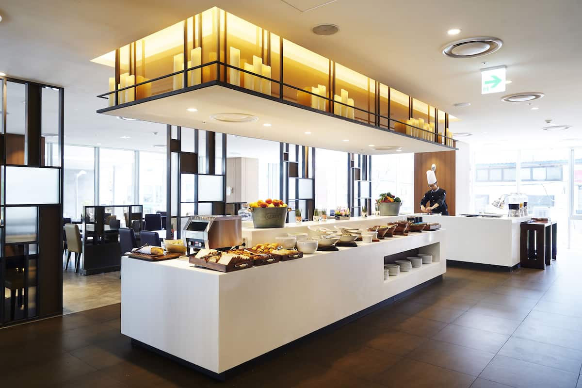 A modern hotel lobby with a central buffet area, seating arrangements, and a large window providing natural light.