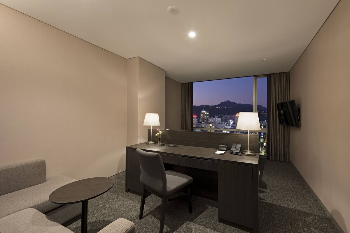 A modern hotel room with a desk, chair, and a view of a cityscape at dusk.