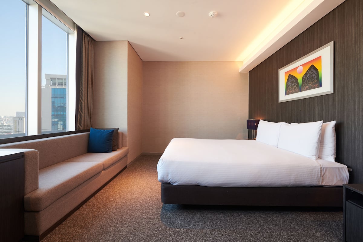 A modern hotel room with a large bed, a sofa, a desk, and a city view.