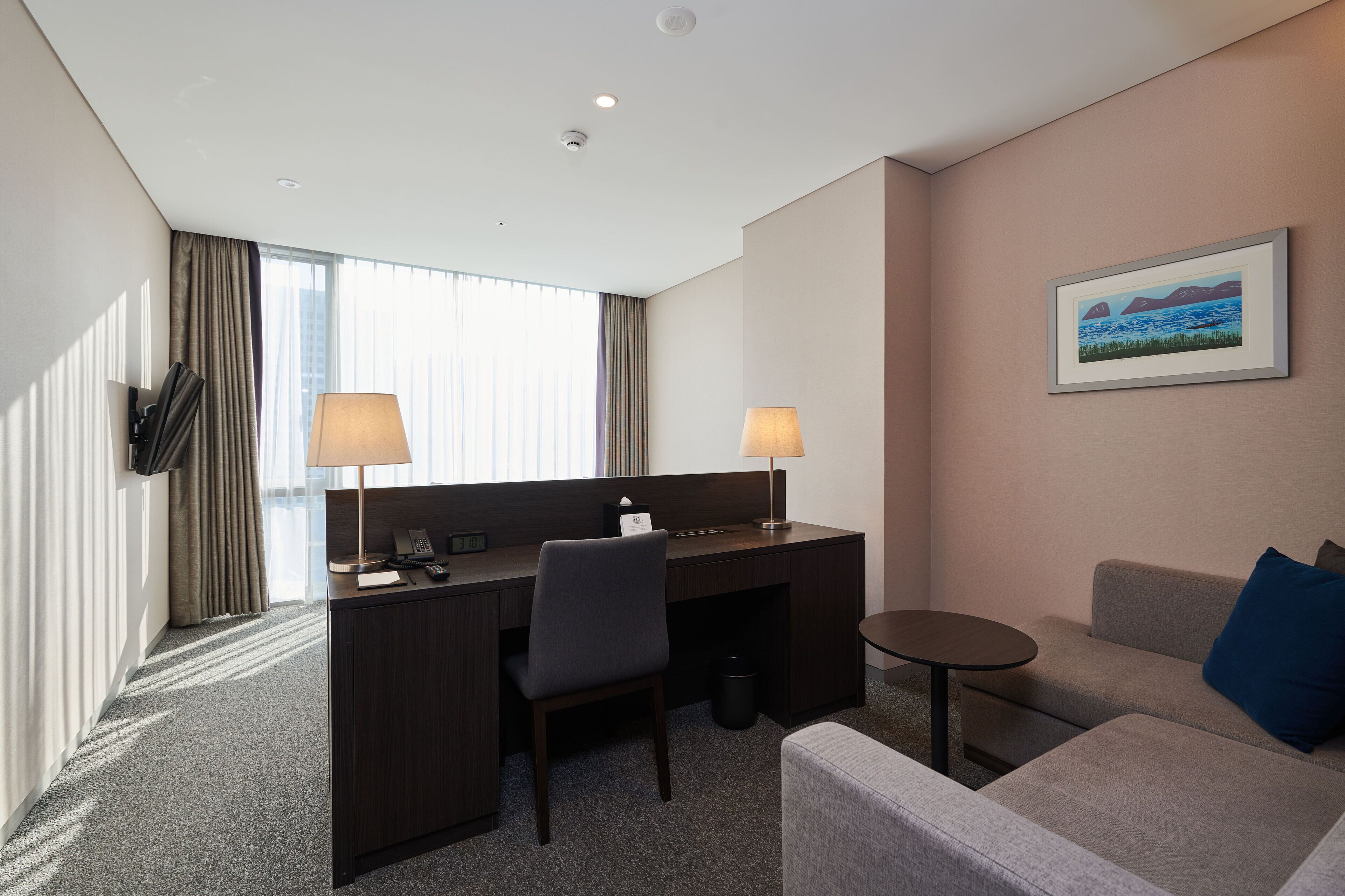 A hotel room with a desk, two chairs, a sofa, a small table, a lamp, a picture on the wall, and a window with curtains.