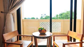 A balcony with two wooden chairs and a small round table, a white curtain, and a view of greenery and a building in the background.
