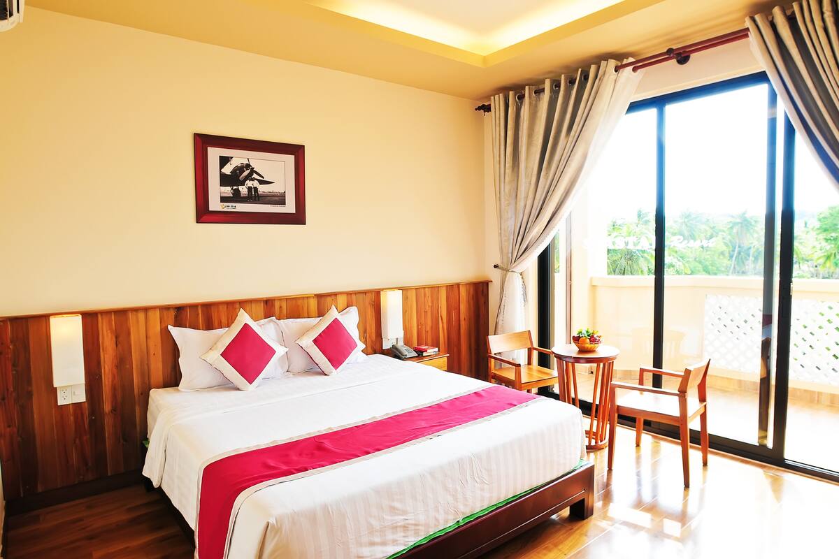 Une chambre d’hôtel avec une tête de lit en bois, un grand lit avec une literie blanche et des touches de rouge, une petite table avec une chaise, et un balcon donnant sur un paysage verdoyant.