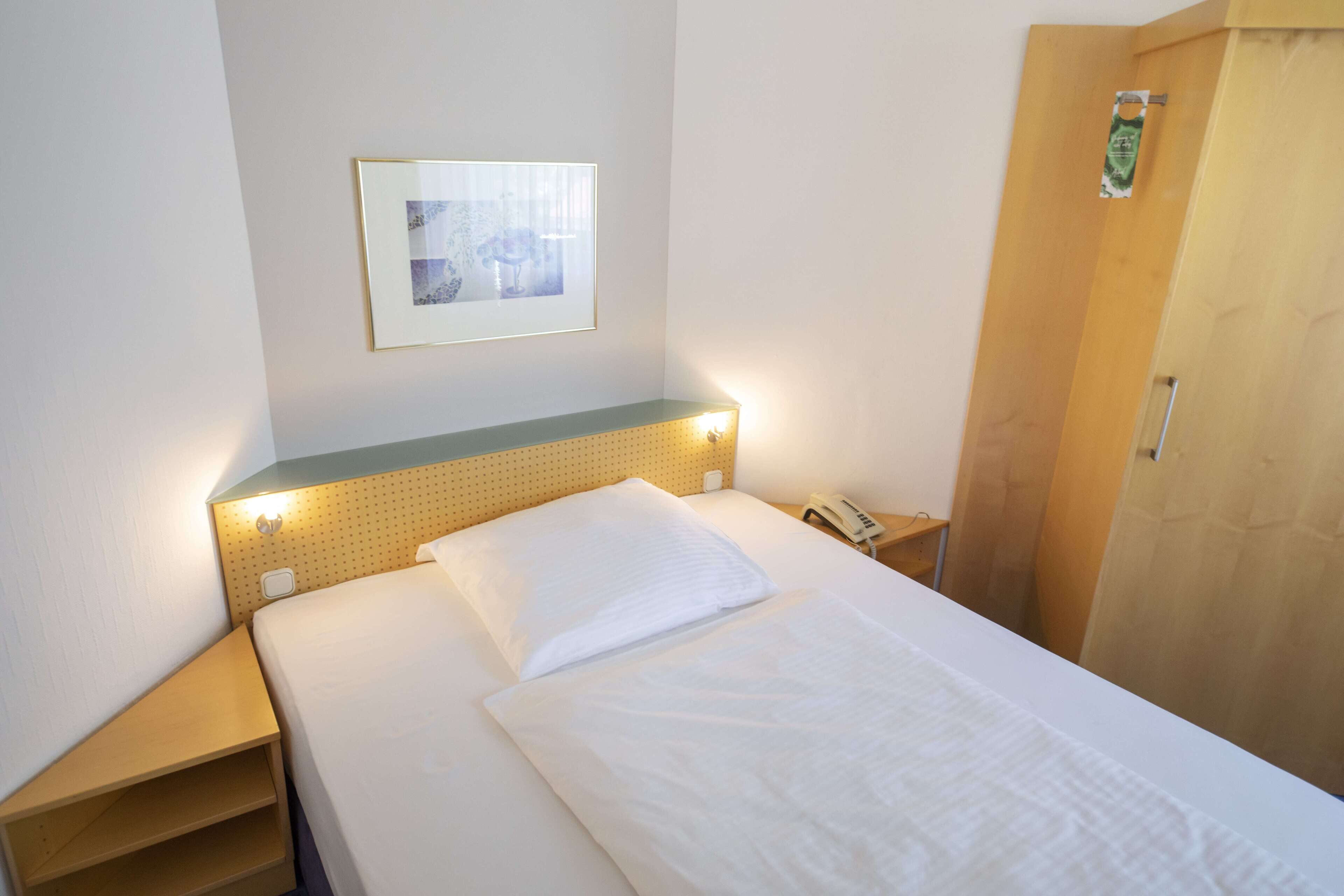 A neatly made bed with a white comforter and pillows, a framed picture on the wall, and a wooden door to the right.