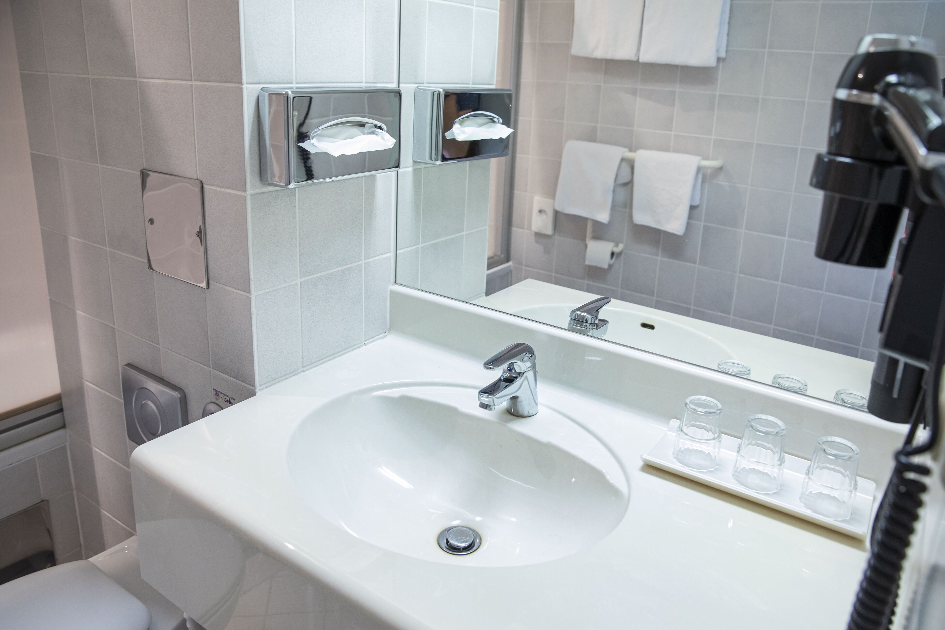 Ein modernes Badezimmer mit einem weißen Waschbecken, einem Chromarmaturen und einem Spiegel darüber. Auf dem Rand des Waschbeckens stehen drei kleine Glasbehälter.