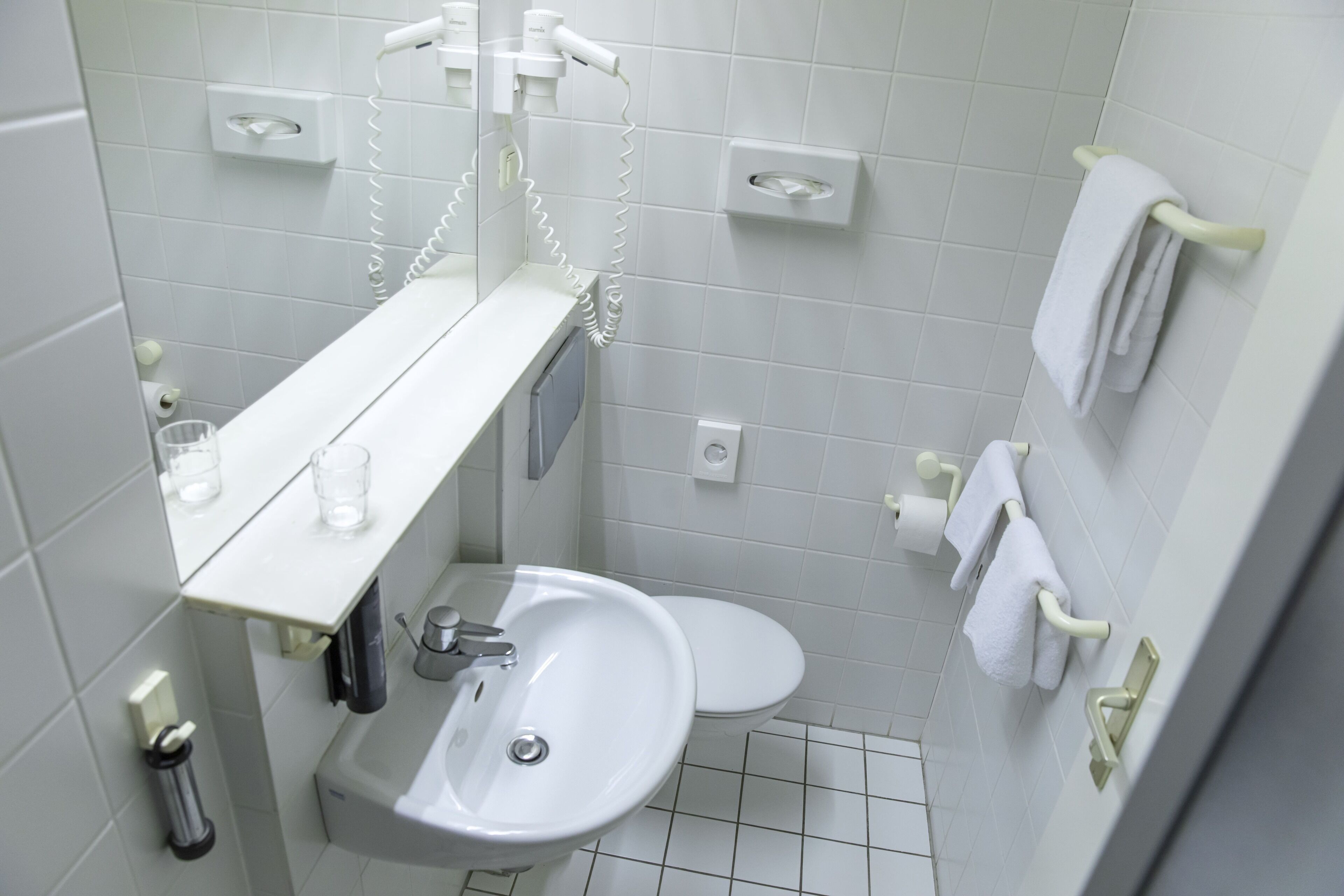 Un baño pequeño y limpio con un lavamanos blanco, inodoro y espejo sobre el lavamanos. Hay dos toalleros en la pared, y una pequeña estantería con una copa y una botella.