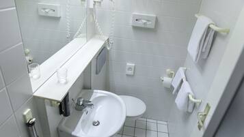 Un baño pequeño y limpio con un lavamanos blanco, inodoro y espejo sobre el lavamanos. Hay dos toalleros en la pared, y una pequeña estantería con una copa y una botella.