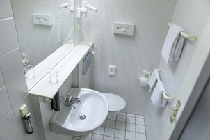 Un baño pequeño y limpio con un lavamanos blanco, inodoro y espejo sobre el lavamanos. Hay dos toalleros en la pared, y una pequeña estantería con una copa y una botella.