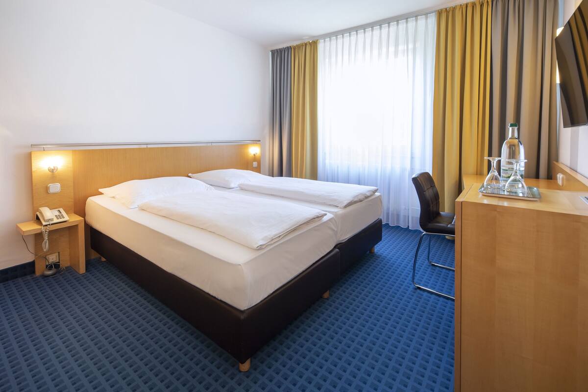 A hotel room with a double bed, a desk with a telephone, a chair, and a large window with curtains.