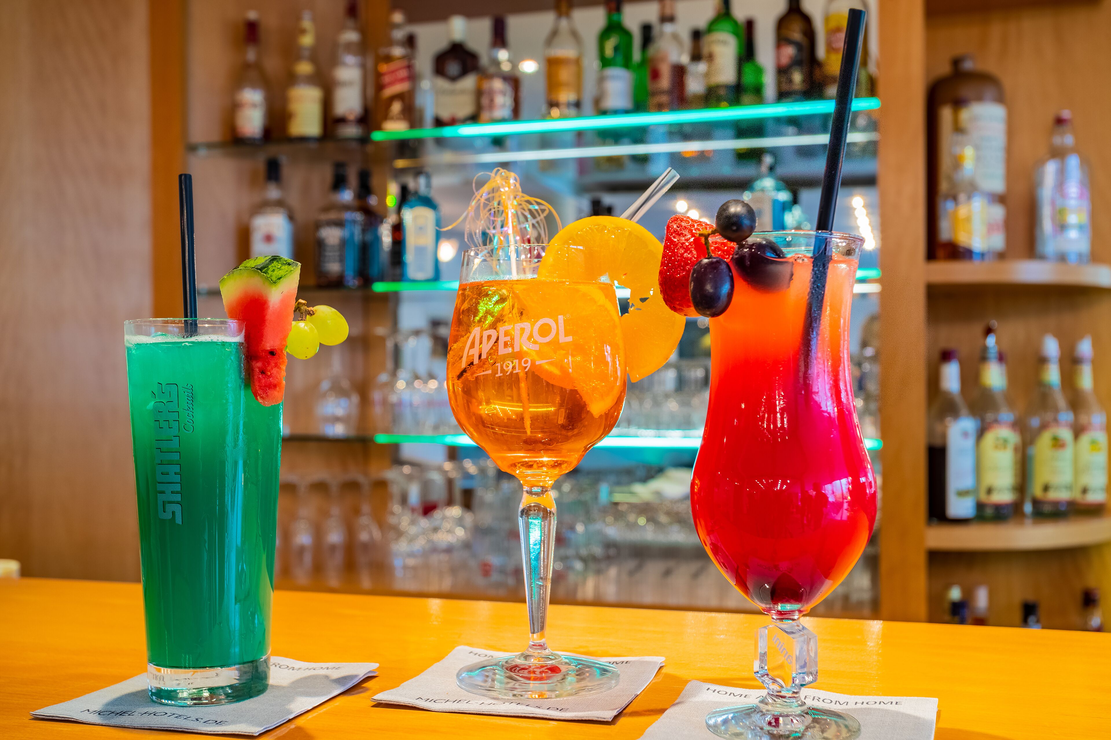 Three cocktails on a bar counter with a background of various bottles.
