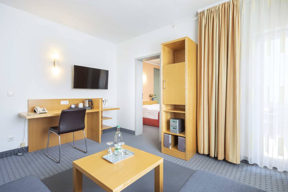 A hotel room with a desk, chair, TV, and a minibar.