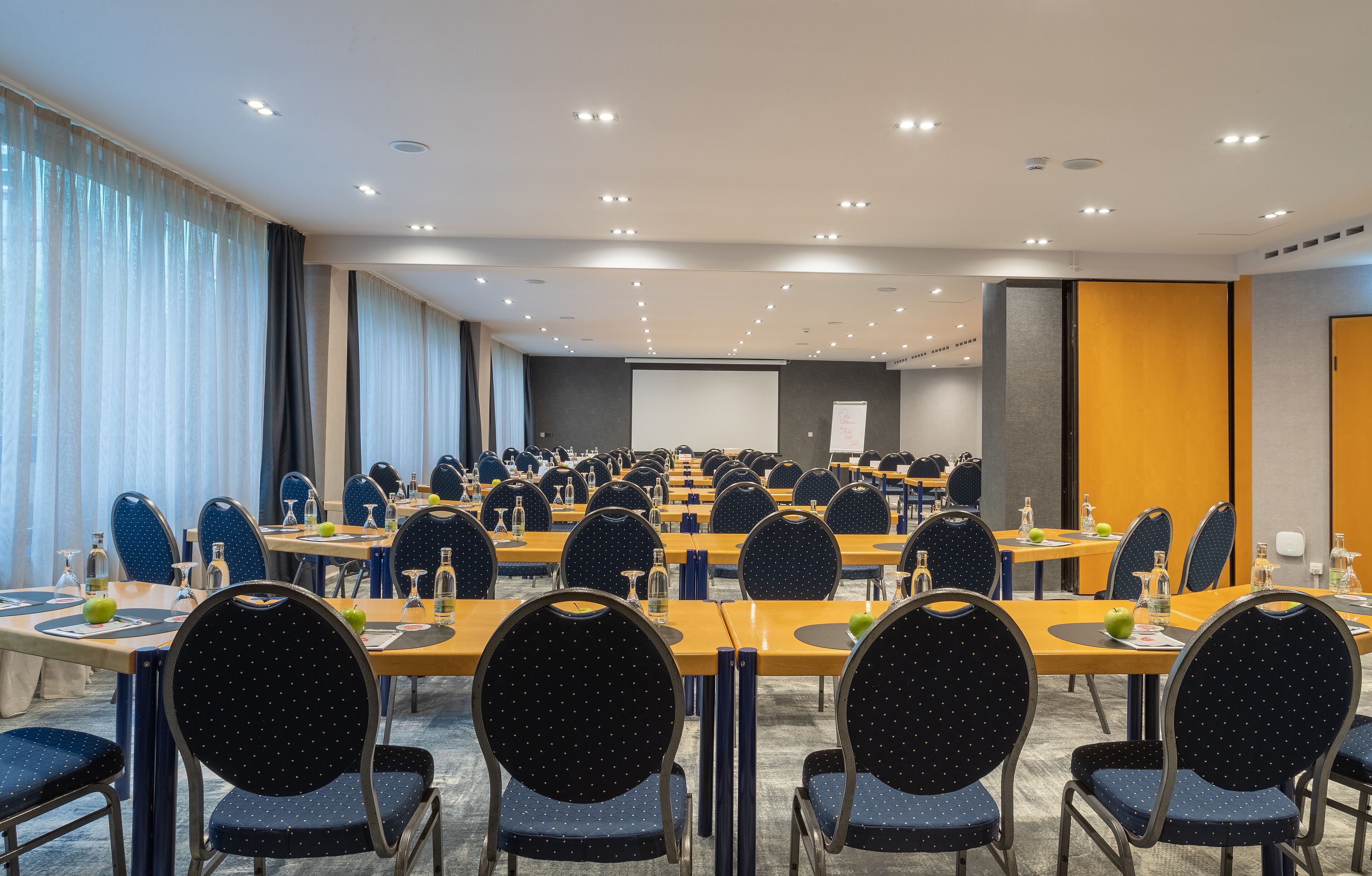 Uma sala de conferências com várias mesas e cadeiras, equipada com placas de identificação, garrafas de água e uma tela de projeção.