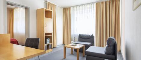 Una habitación de hotel moderna con un escritorio de madera, un sofá gris, una mesita pequeña y una estantería.