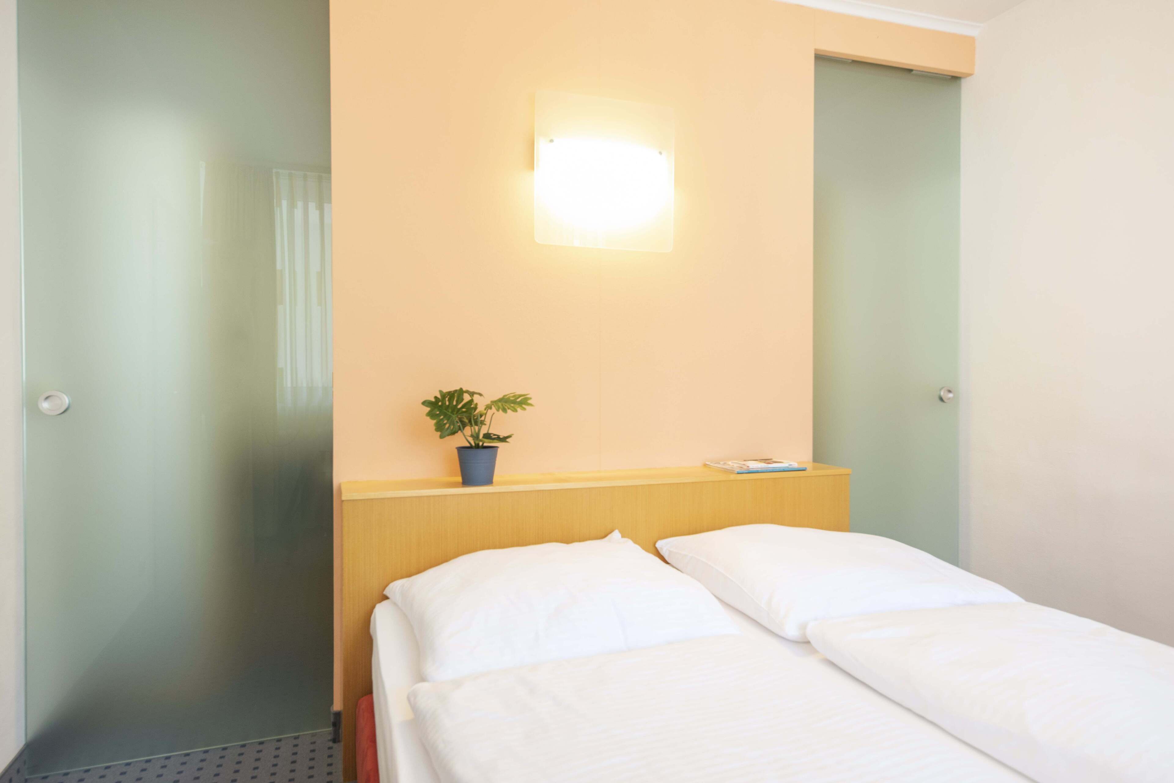 Una camera d'hotel con un letto, un comodino, una pianta in vaso e un punto luce a parete.